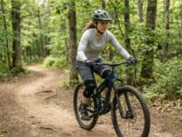 A beginner mountain biker wearing a helmet, knee pads, and gloves on a green trail